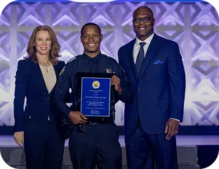 Three people standing in front of a purple geometric patterned wall, with the center person holding a framed award plaque and smiling.