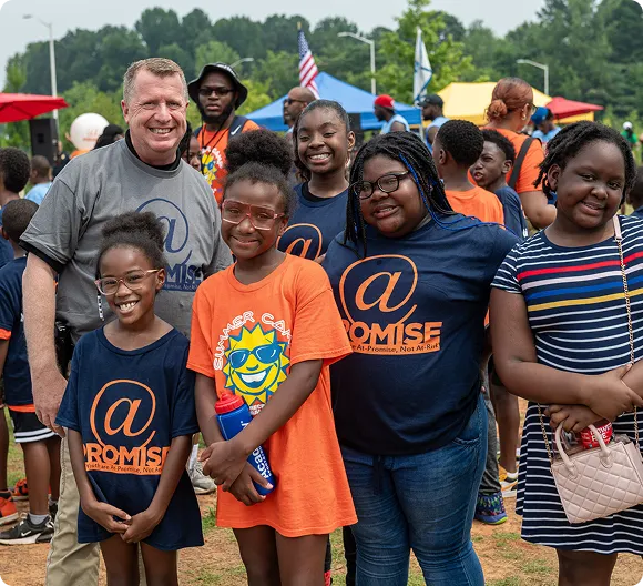 Smiling group of diverse children and one adult at an outdoor event with tents and flags in the background.