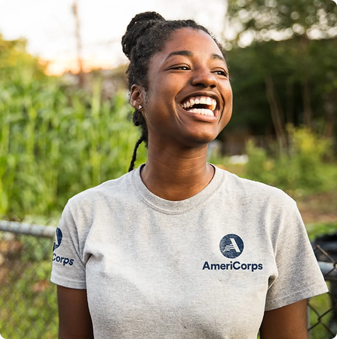 Smiling young woman wearing a gray AmeriCorps t-shirt standing outdoors in a garden.