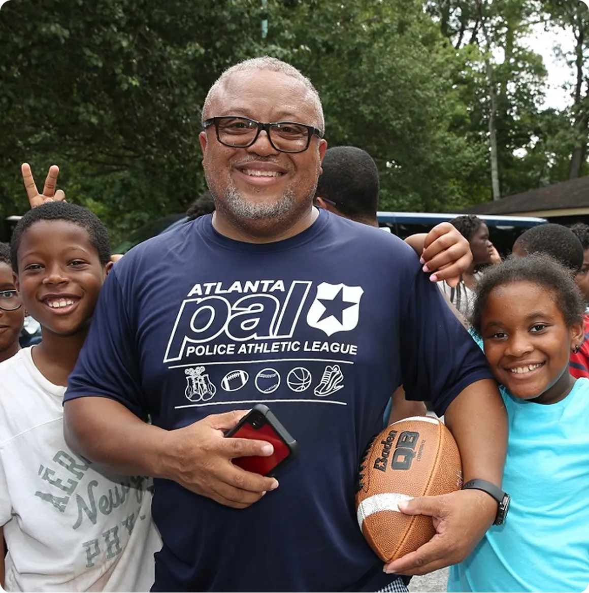 Smiling man in glasses and Atlanta PAL shirt holding a football, flanked by two happy children outdoors.