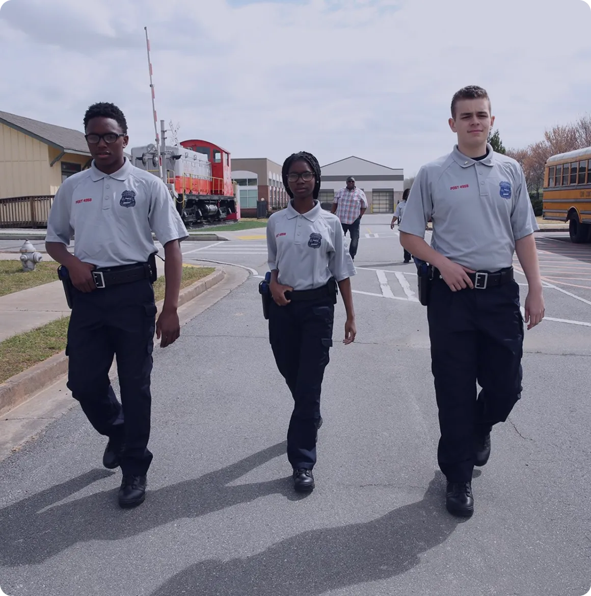 Three young cadets in uniform walking confidently on a street with a red train and school bus in the background.
