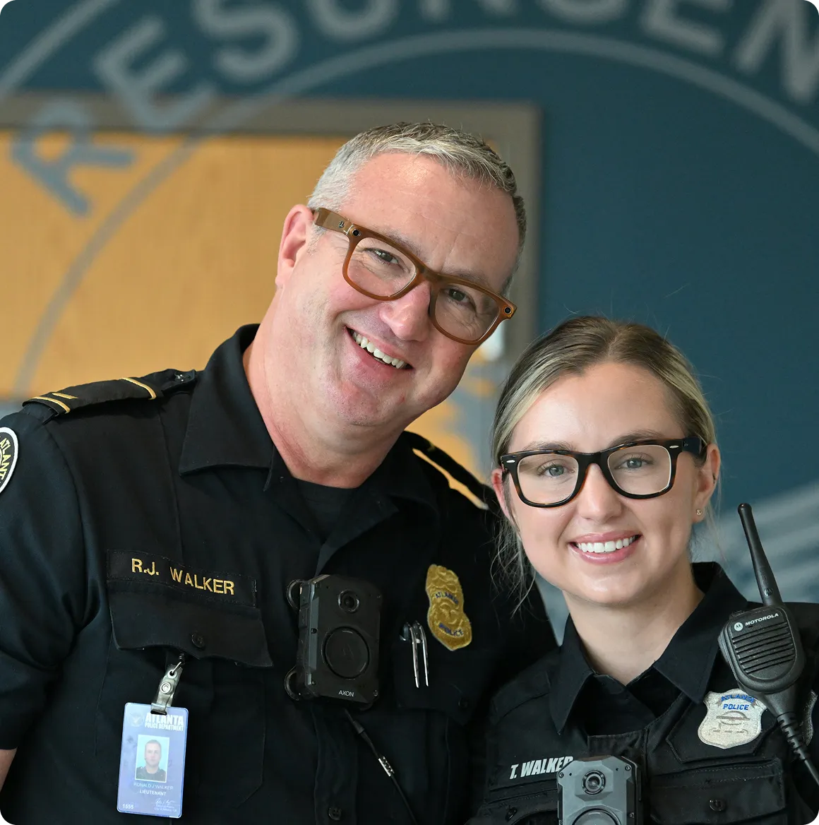 Two smiling police officers wearing glasses and uniforms with body cameras and radios.