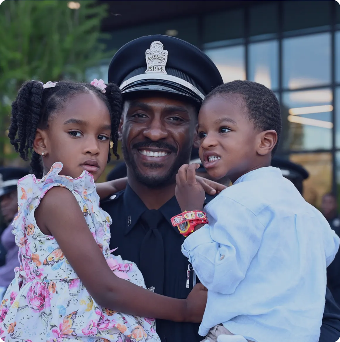 Smiling African American police officer holding two young children outdoors near a building.