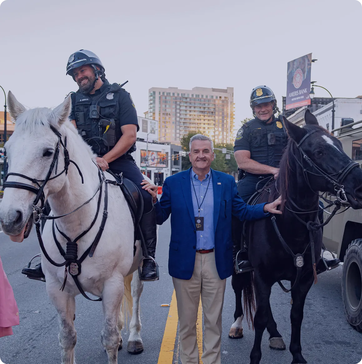 Two mounted police officers smiling on horses with a man standing between them on a city street.