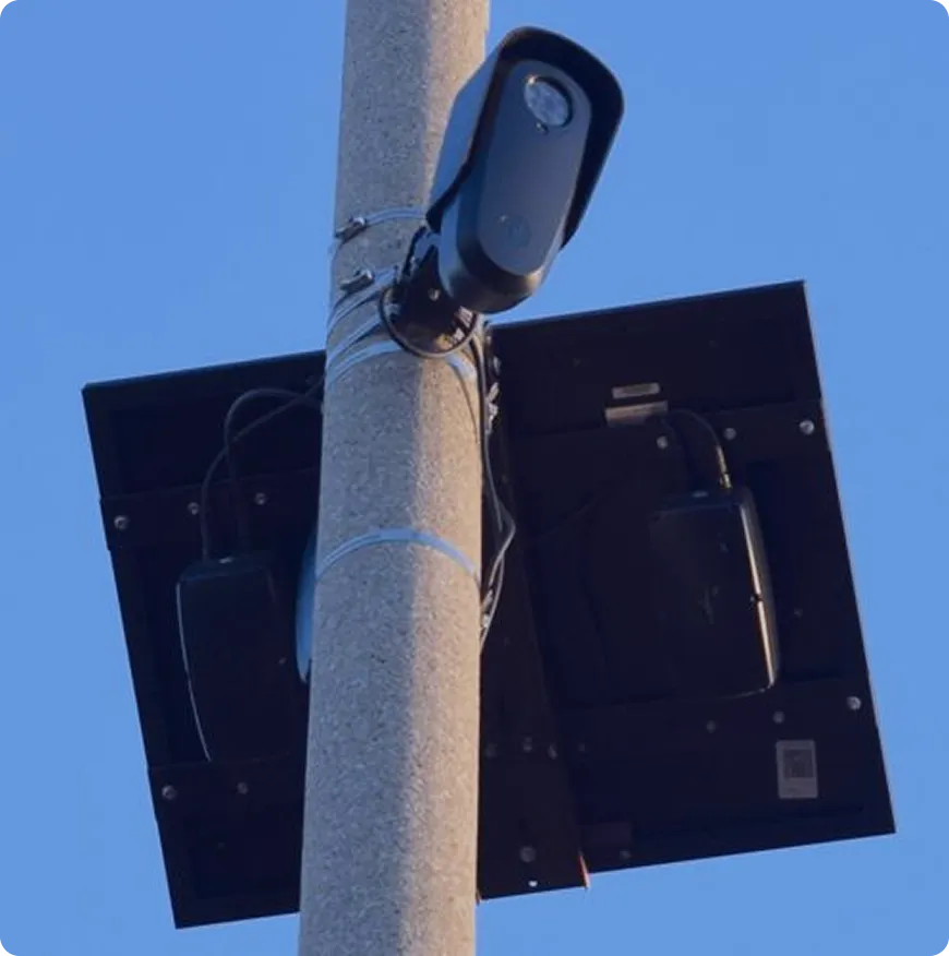 Surveillance camera mounted on a pole with a solar panel attached behind it against a clear blue sky.