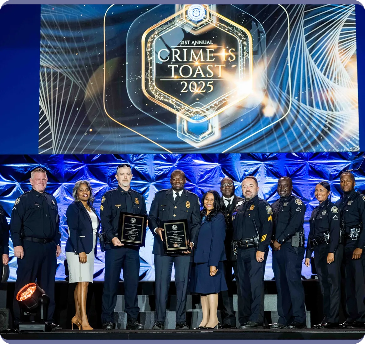 Group of police officers and two women in formal attire standing on stage with plaques during the 21st Annual Crime Stoppers Toast 2025 event.