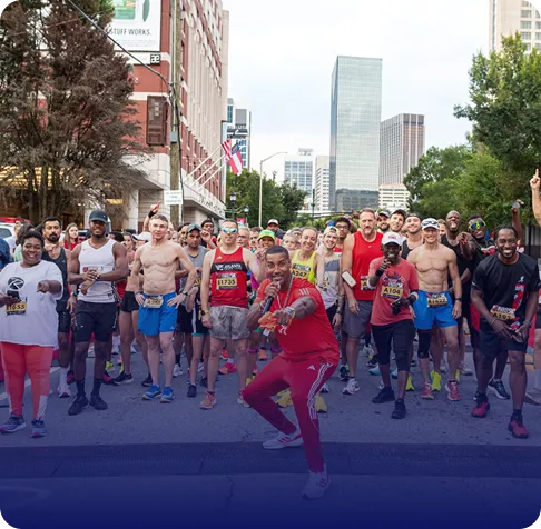 Group of diverse runners gathered at the starting line of a city road race, with a man in red in front pointing at the camera.