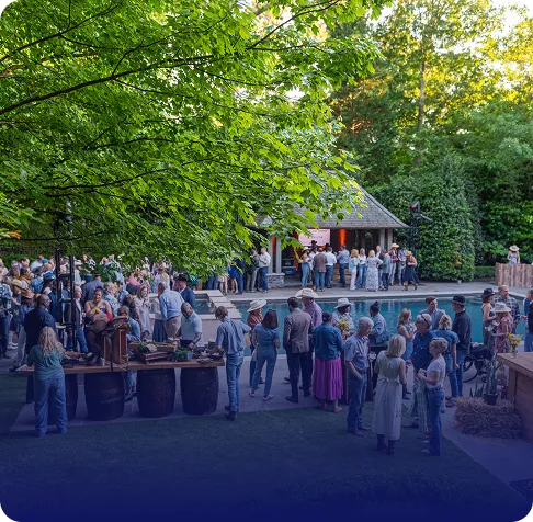 Crowd of people socializing around a pool near a small pavilion in a shaded outdoor park setting with green trees.