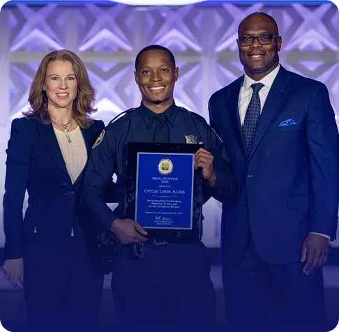 Three people in formal attire smiling, with the center person in a police uniform holding an award plaque.