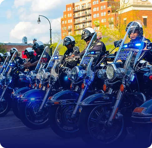 Line of police officers wearing helmets sitting on motorcycles with flashing blue lights in an urban setting.