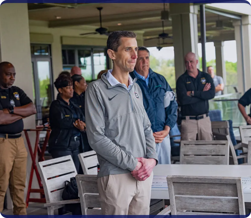 A group of people standing indoors, with one man in a grey Nike pullover and khaki pants in the foreground, and several others, some in uniform, in the background.