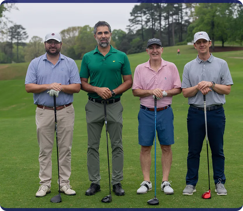 Four men standing side by side on a golf course, each holding a golf club and dressed in casual golf attire.