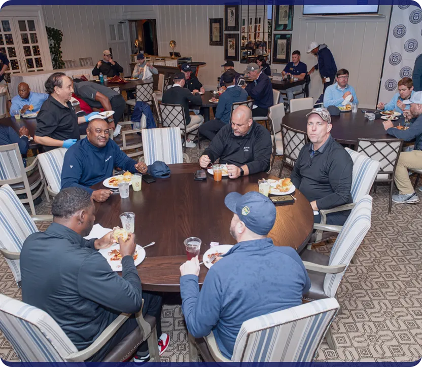 Group of men gathered around multiple tables eating and conversing in a spacious dining room.