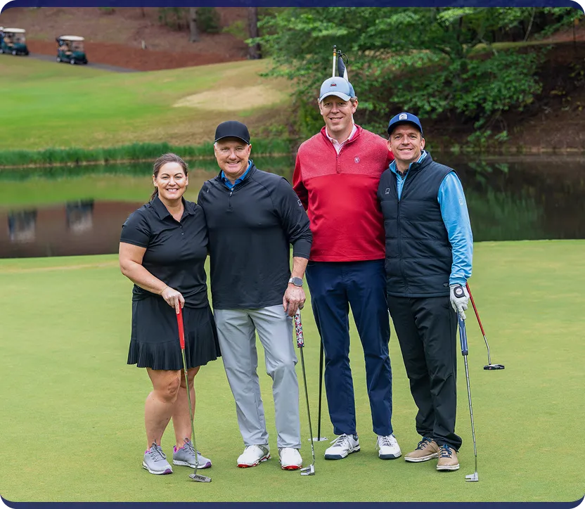 Four people standing together on a golf course green, each holding a putter, with a pond and golf carts in the background.