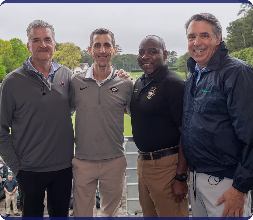 Four men standing together and smiling outdoors with a golf course in the background.