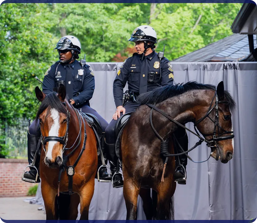 Two police officers wearing helmets and uniforms mounted on brown horses outdoors.