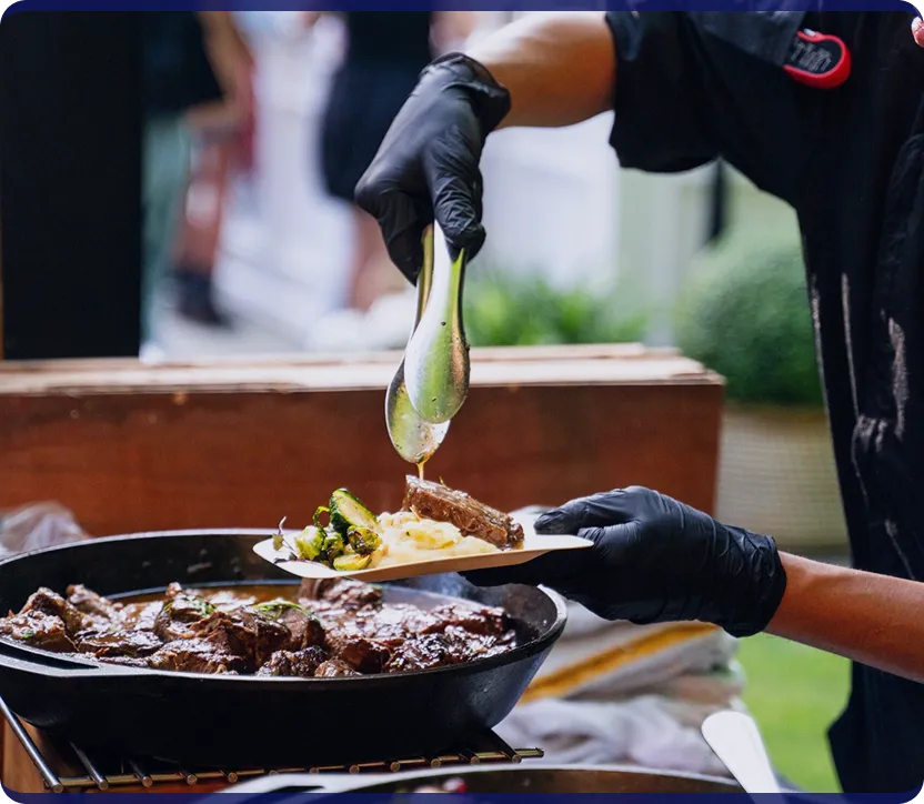 Person wearing black gloves serving cooked meat and vegetables onto a plate using tongs from a pan.