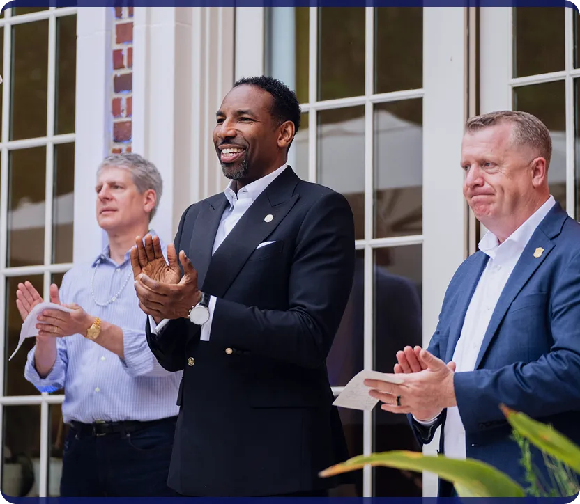 Three professionally dressed men standing outdoors clapping during an event.