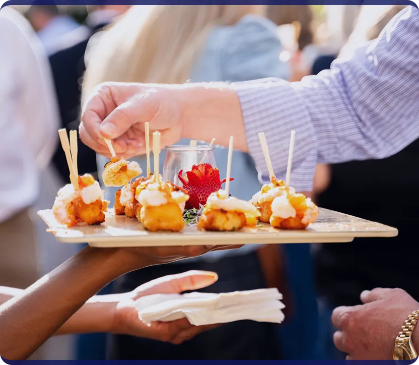 Person picking up a shrimp appetizer with a stick from a serving tray decorated with a carved strawberry flower.
