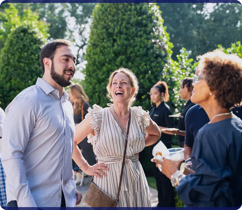 Three adults smiling and talking outdoors at a social gathering with green bushes in the background.