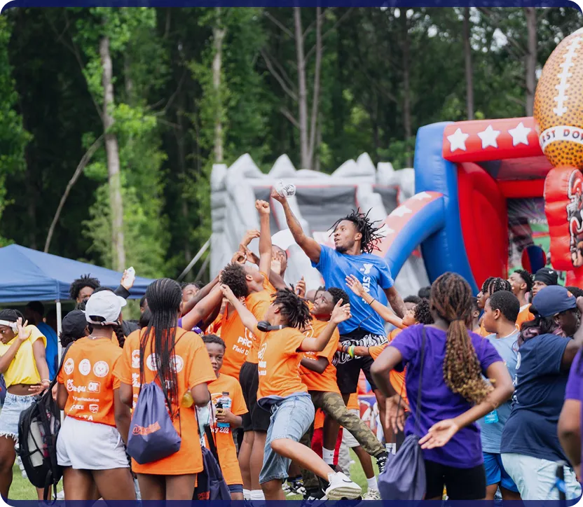 Group of children and teens playing and reaching for a ball during an outdoor event with inflatable play structures in the background.