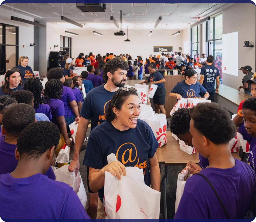 Volunteers in navy shirts handing out Chick-fil-A bags to children in purple shirts inside a bright room.