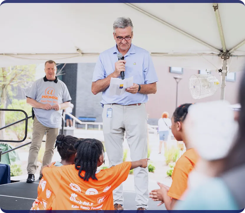 Man with gray hair and glasses speaking into a microphone while reading from a paper under a tent at an outdoor event, with children in orange shirts in the foreground.
