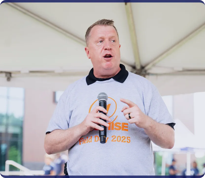 Man speaking into a microphone while wearing a gray t-shirt that says 'Promise Field Day 2025' under a tent.