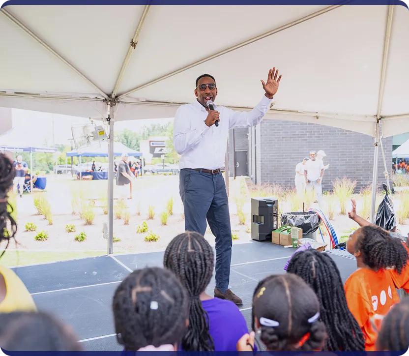 Man in white shirt and sunglasses speaking into a microphone on a stage under a tent, engaging with seated children facing him.