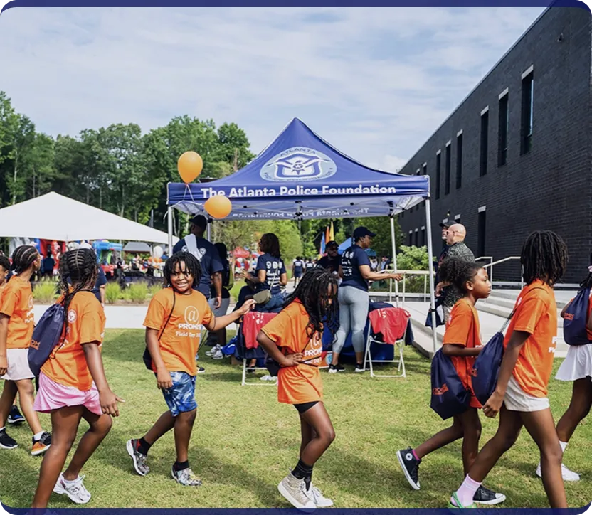 Group of children in orange shirts walking outdoors near a tent with The Atlanta Police Foundation logo during a community event.