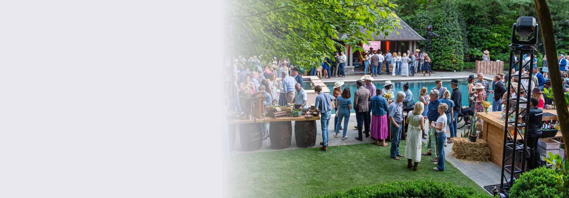 Large outdoor gathering of people socializing near a pool with a bar setup and greenery around.