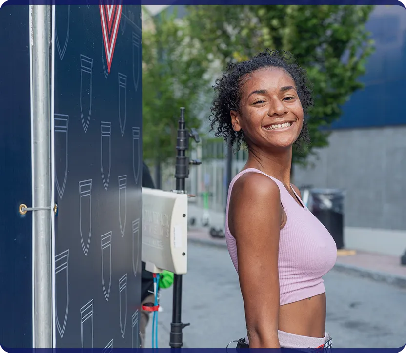 Smiling young woman with curly hair wearing a pink crop top standing outdoors near equipment and trees.