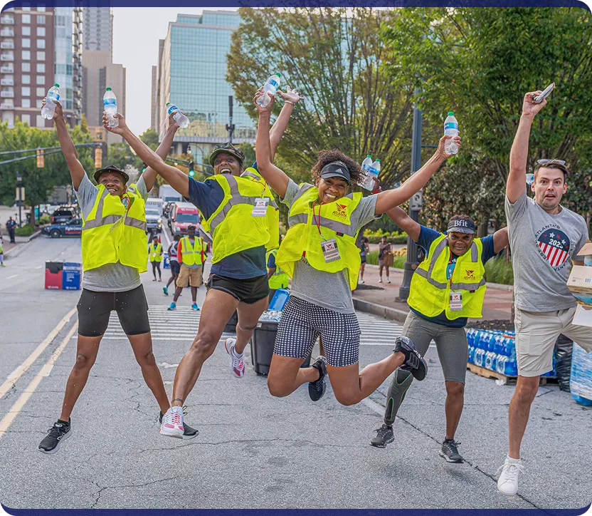 Five people wearing yellow safety vests joyfully jumping in the middle of a city street, holding water bottles.