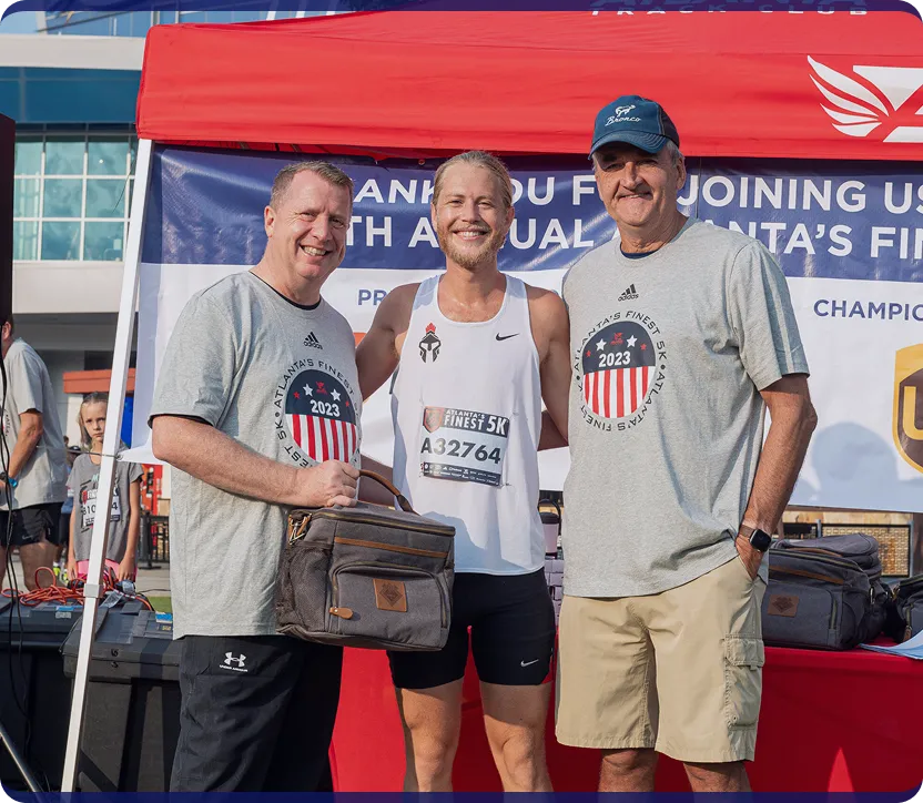 Three men smiling under a red tent at Atlanta's Finest 5K event, with two wearing grey event t-shirts and one in white running gear holding a bag.