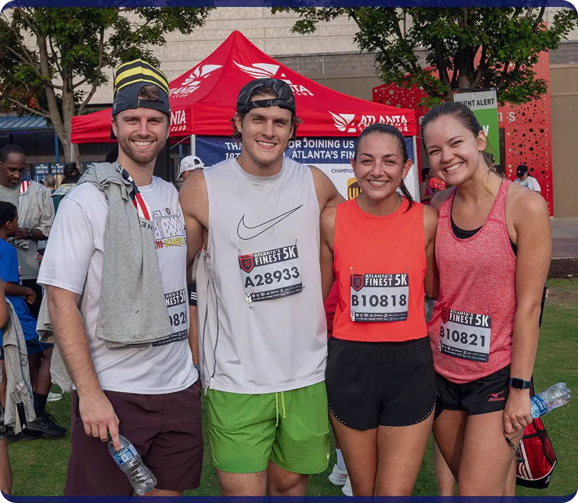 Four smiling runners posing outdoors with race bibs for Atlanta's Finest 5K in front of a red event tent.