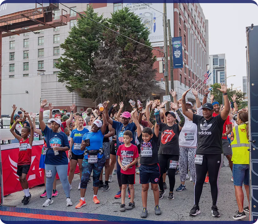 Group of diverse runners, including children and adults, standing at a race start line with hands raised in excitement during a city running event.
