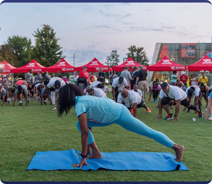 A woman in blue athletic wear leads a group stretching on grass at an outdoor event with red Atlanta tents in the background.