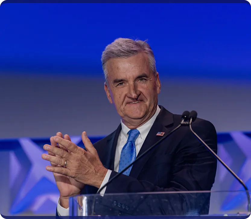 Man in a dark suit with an American flag pin clapping hands behind a transparent podium with microphone.