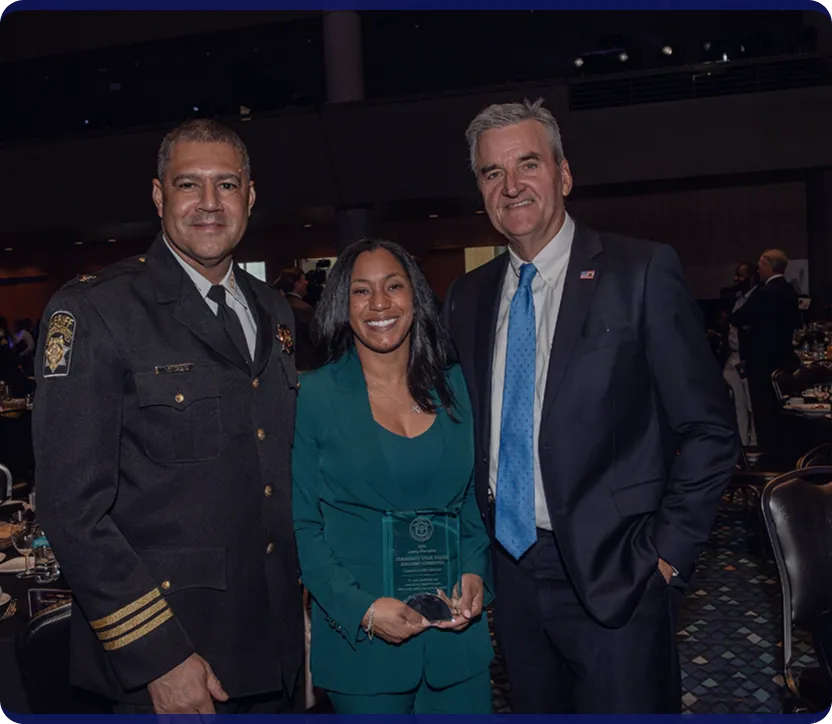 Three individuals smiling at an event, with the woman in the middle holding an award plaque; a man in police uniform on the left and a man in a suit with a blue tie on the right.