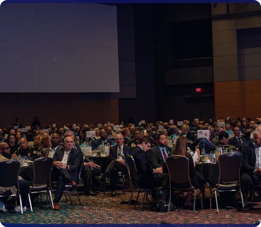 Large group of professionally dressed people seated at round tables in a dimly lit conference or banquet hall.