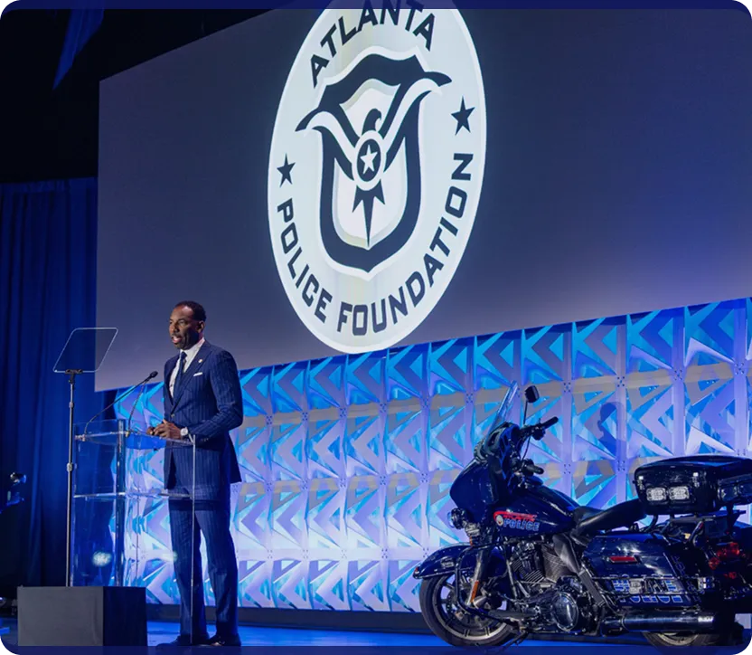 Man in a blue suit speaking at a clear podium with Atlanta Police Foundation logo projected behind and a police motorcycle on stage.