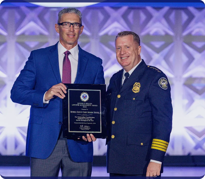 Two men standing side by side; one in a blue suit holding a plaque, and the other in a police dress uniform smiling.