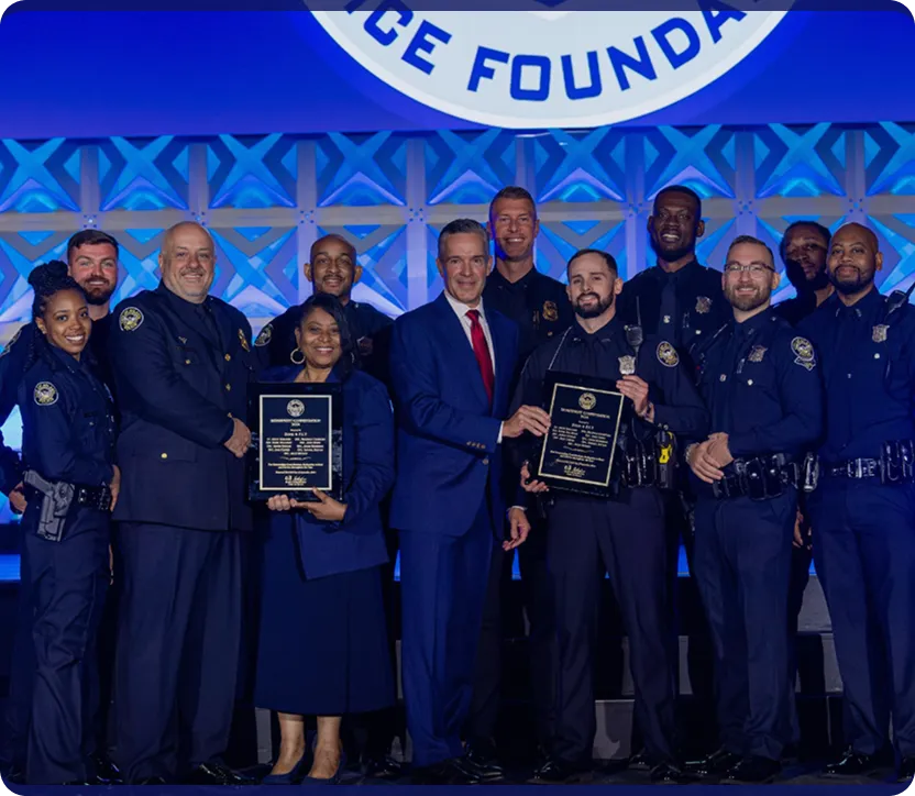 Group of police officers and a man in a suit standing on stage, two holding plaques during an award ceremony.