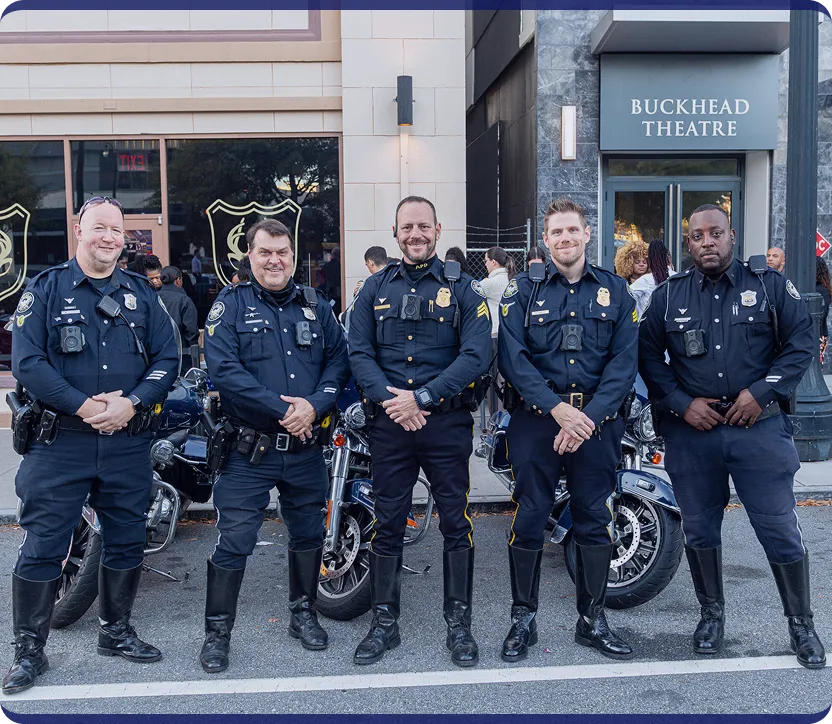 Five uniformed police officers standing in front of motorcycles on a street with a Buckhead Theatre sign in the background.