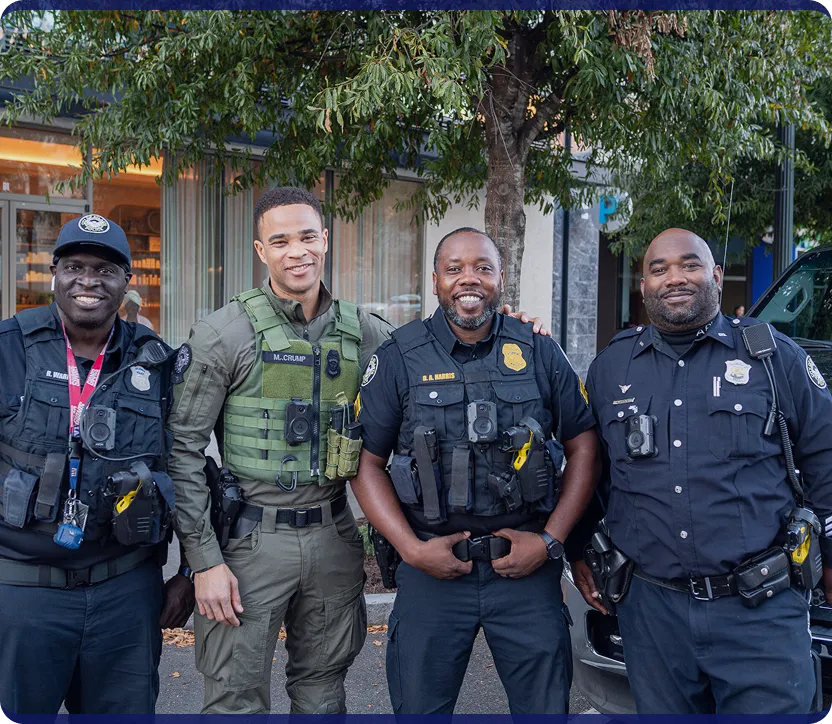 Four uniformed police officers standing together outside with a tree and building in the background, smiling at the camera.