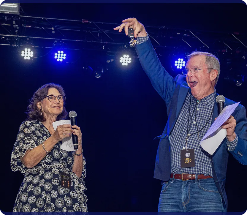 A man and woman on stage holding microphones and papers; the man is raising a set of keys enthusiastically.