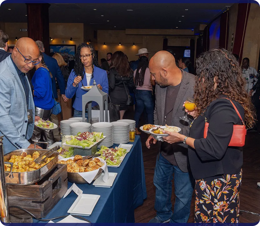 People serving themselves food from a buffet table with plates, croissants, salad, and roasted potatoes at an indoor event.