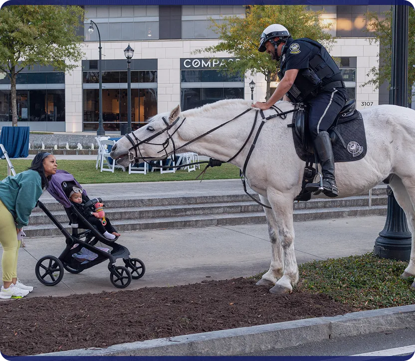 Police officer on a white horse leaning forward to interact with a baby in a stroller pushed by a woman on a city sidewalk.