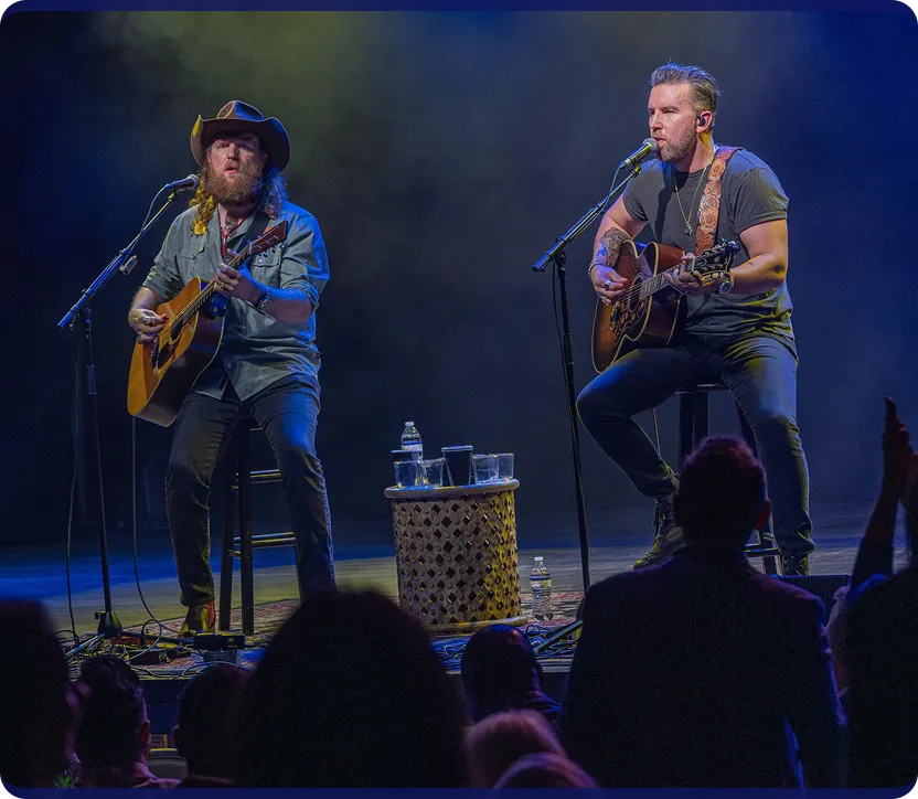 Two male musicians performing an acoustic set on stage, each playing an acoustic guitar and singing into microphones, with a small table holding drinks between them and an audience visible in the foreground.