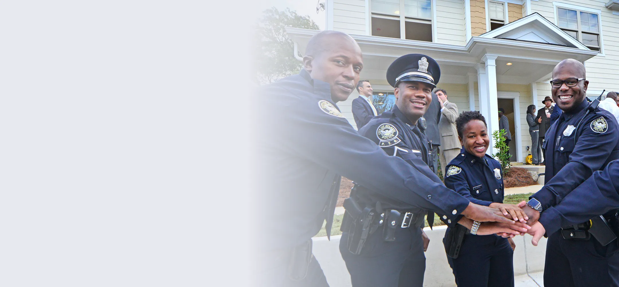 Four smiling police officers in uniform joining hands in front of a house with people standing in the background.
